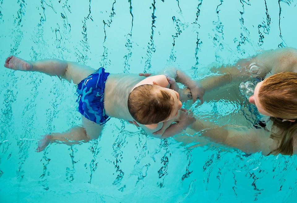 Liten kille står på kanten och ska hoppa i bassängen till mamma som står i vattnet.