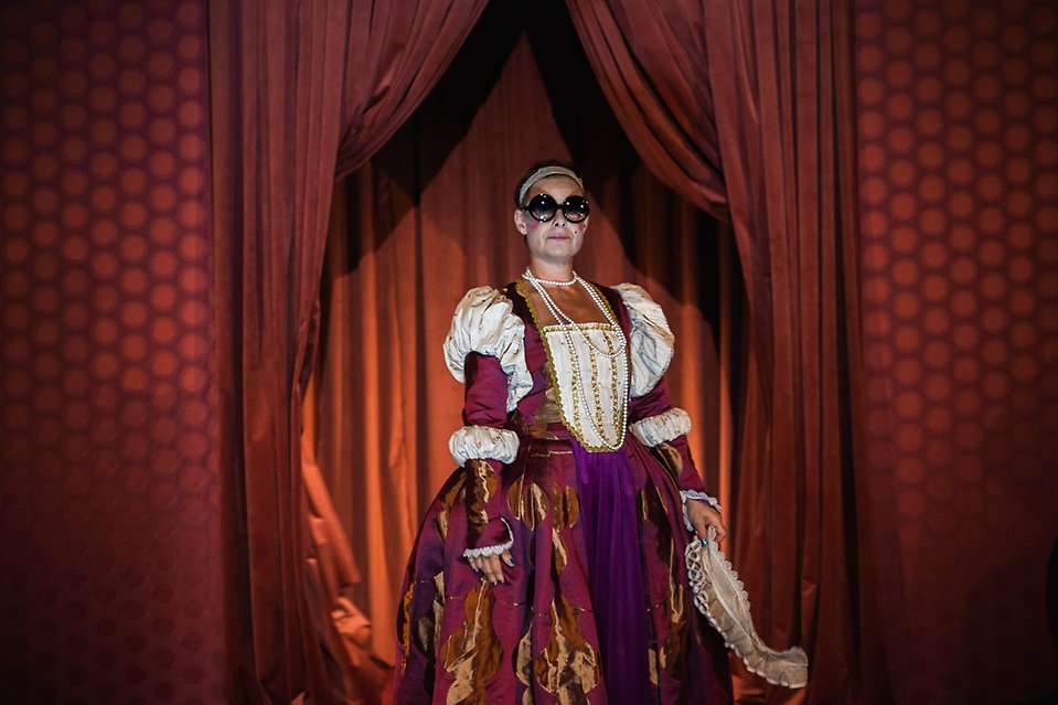 Foto från föreställningen "Jag är drottning" på en person i 1600-tals kläder med solglasögon som står framför en röd ridå. 
