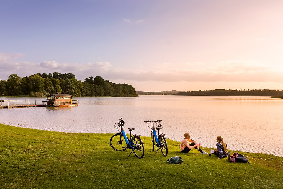 Bild på två ungdomar med cyklar som sitter på gräset intill strandkanten vid Sövdesjön.
