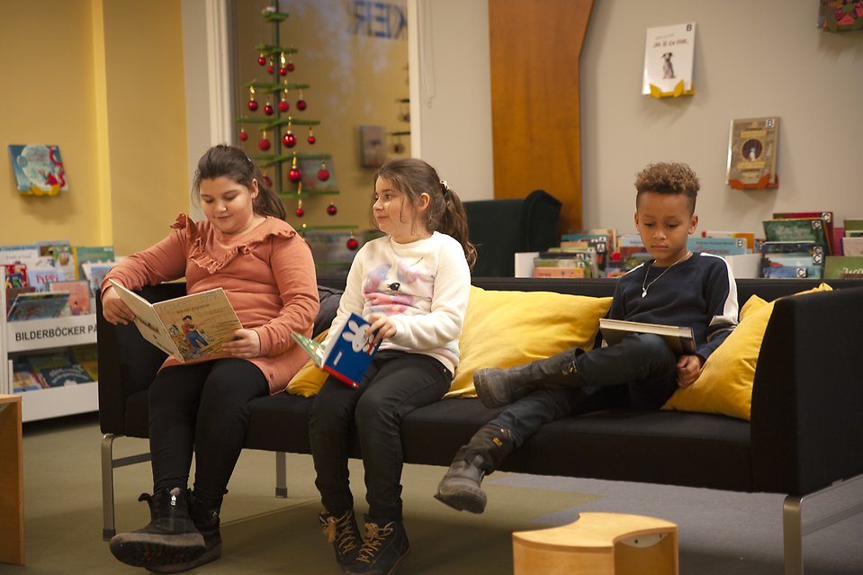 Tre barn sitter i en soffa på barnavdelningen i Sjöbo bibliotek och läser varsin bok