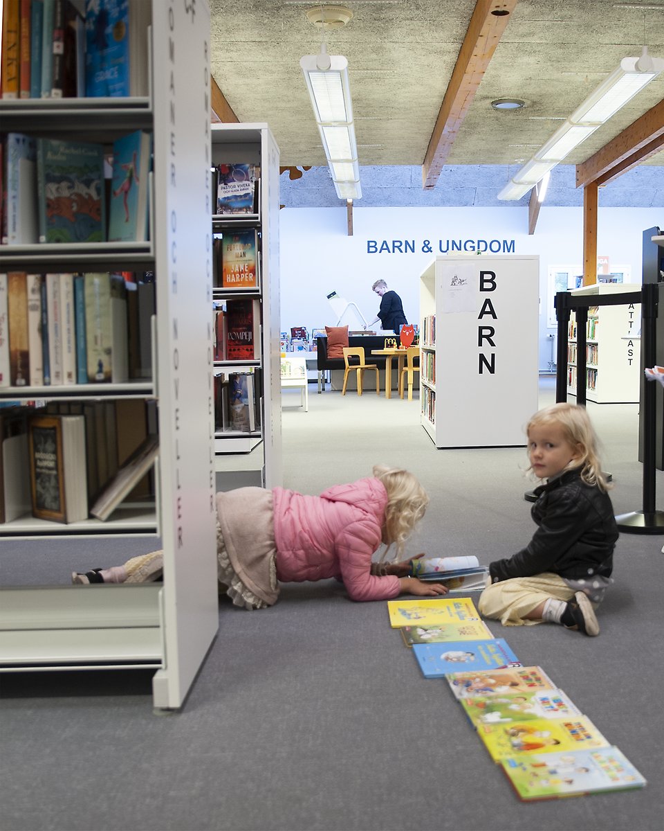 Interiör från Sjöbo bibliotek. Man ser hyllor med böcker, en hylla med texten Barn i bakgrunden samt två barn som har lagt ut böcker på golvet i en lång rad.