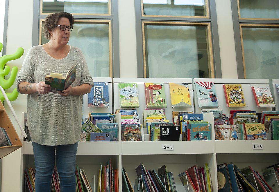 Interiör från Vollsjö bibliotek. En bibliotekarie står framför de låga barnbokhyllorna och håller i en bok