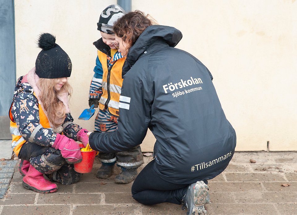 En kvinna i svart jacka sitter på huk och leker med två barn i utekläder. Kvinnan har en svart jacka med texten "Förskolan Sjöbo kommun #tillsammans".
