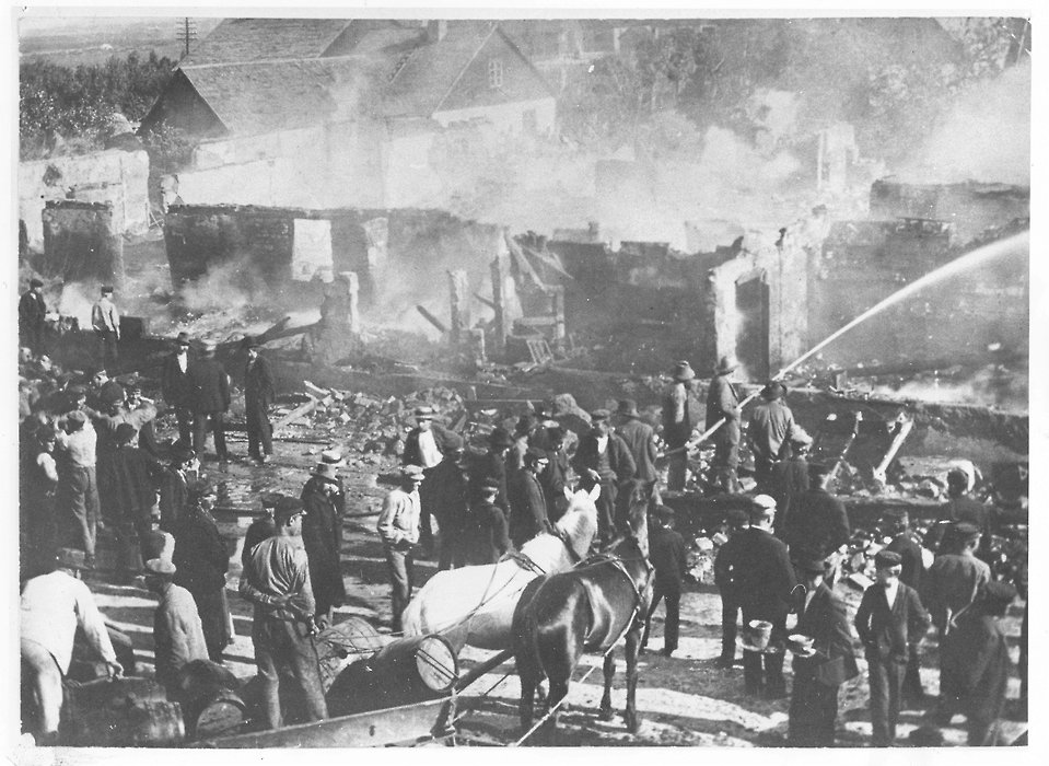 En bild på en bondgård i lågor, med brandkåren (hästdragen kärra, brandslang och handpump) som försöker släcka elden. 